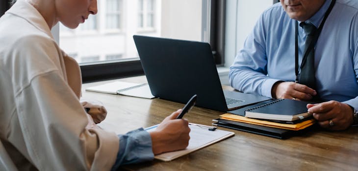 Two professionals engaged in a business meeting, discussing documents with a laptop at the office.
